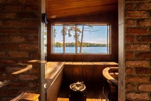 A wooden cabin interior with a large window overlooking a lake and trees, built-in benches, and a rustic stone wall. - Stay North - Vuohilahti (Valkeakoski)