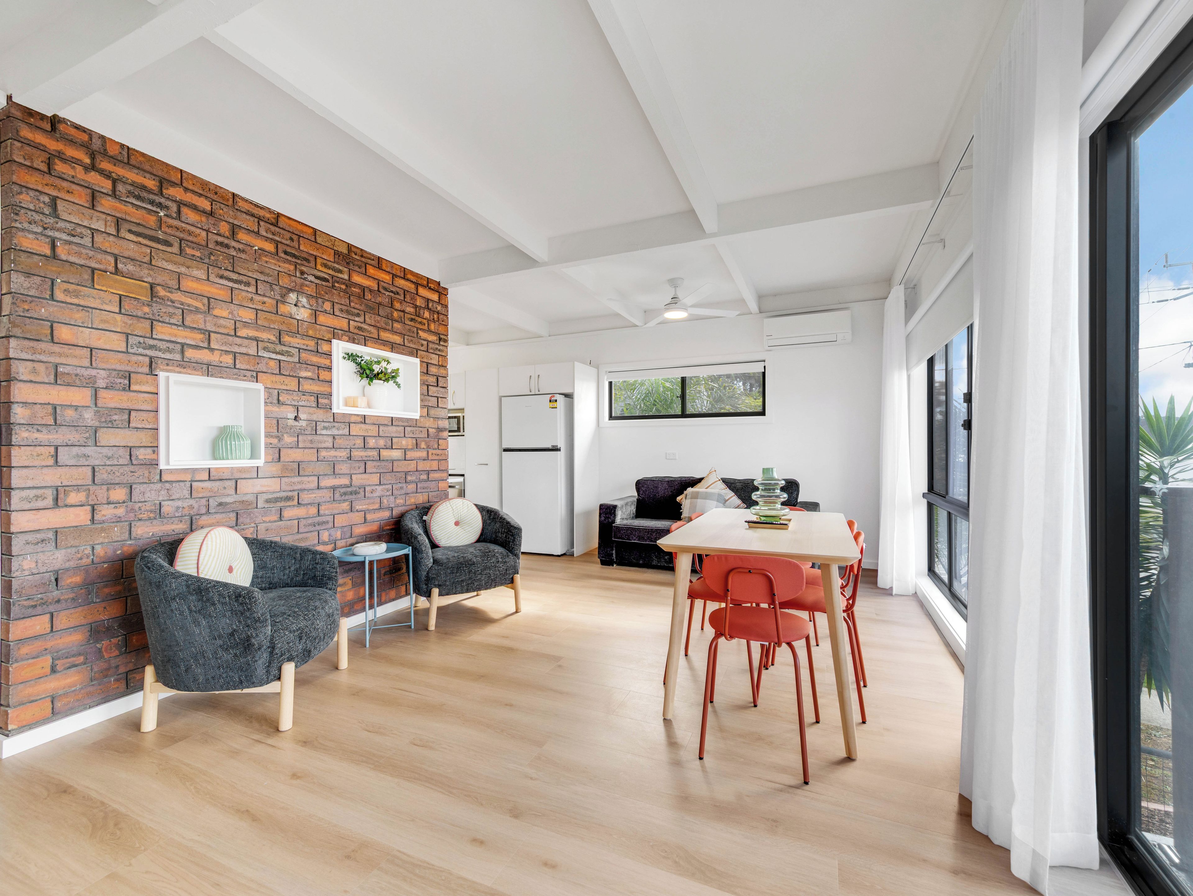 A modern interior with a brick wall, a dining area with red chairs, and a kitchen with white cabinets.