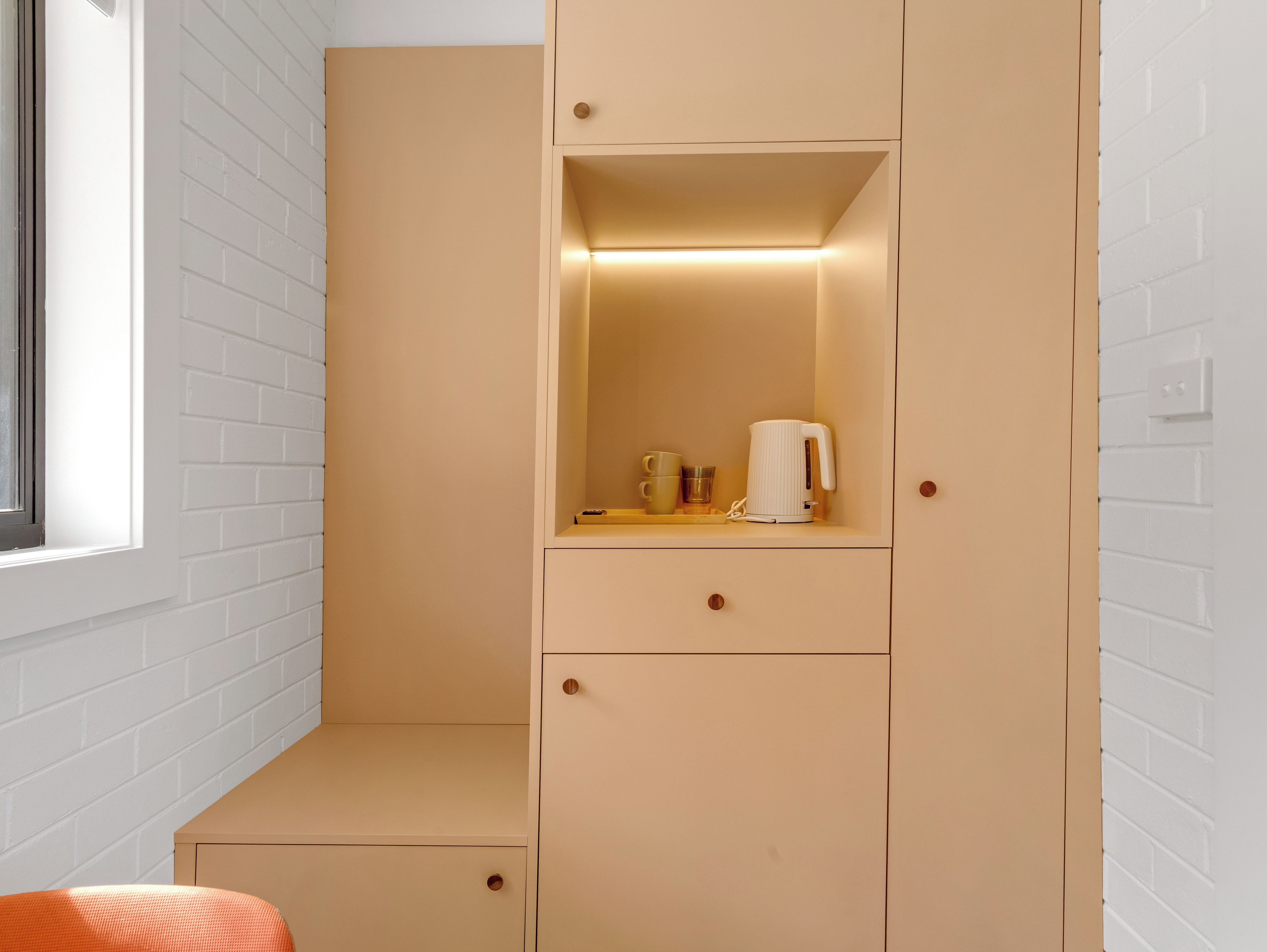 Ein Zimmer mit einer weißen Ziegelwand, einem beige gefärbten Schrank mit Schubladen, einem kleinen Tisch mit Wasserkocher und Tassen sowie einem Fenster.
