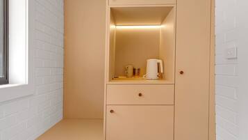 Ein Zimmer mit einer weißen Ziegelwand, einem beige gefärbten Schrank mit Schubladen, einem kleinen Tisch mit Wasserkocher und Tassen sowie einem Fenster.