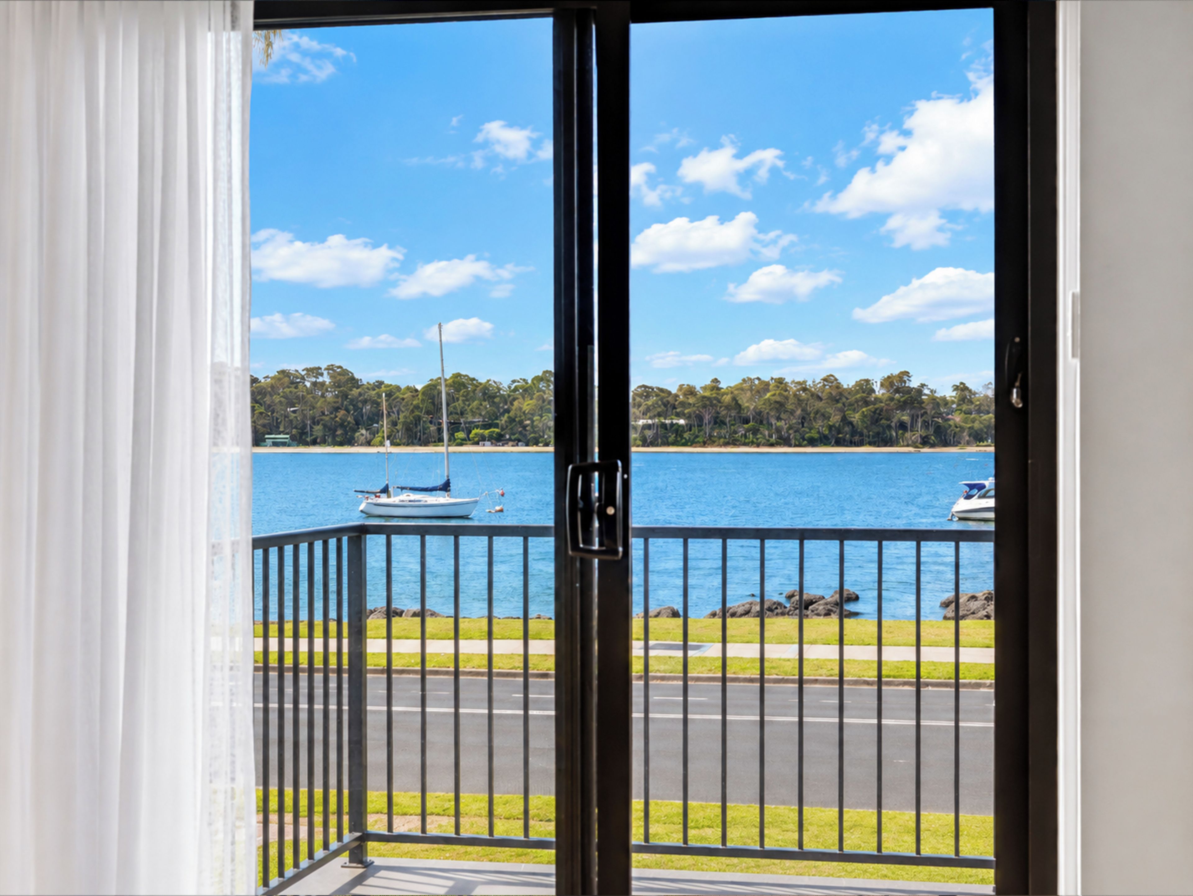 A view from a balcony with a railing, overlooking a body of water with boats, a beach, and a forested area.