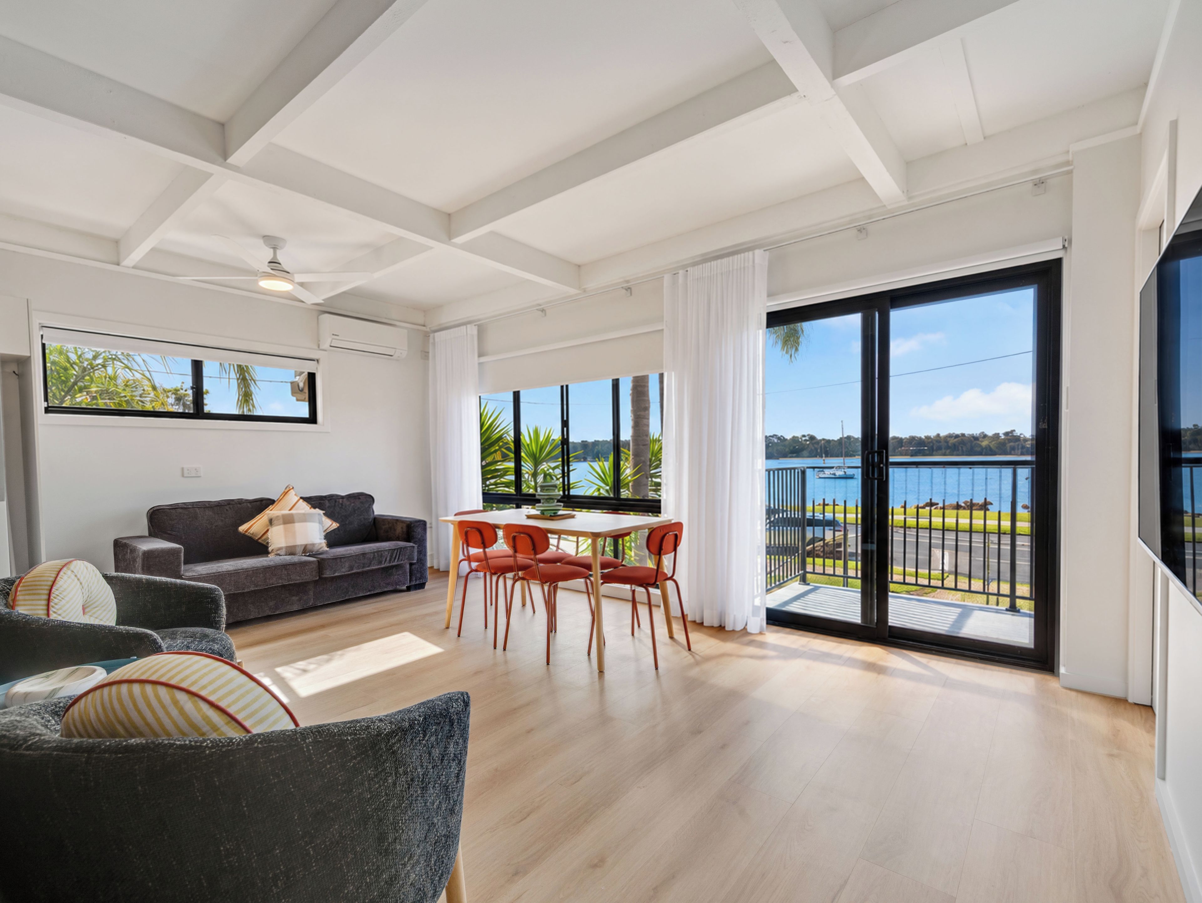 A modern living room with a sofa, dining table, and chairs, large windows with a view of the water, and a balcony with a railing.