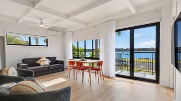A modern living room with a sofa, dining table, and chairs, large windows with a view of the water, and a balcony with a railing.