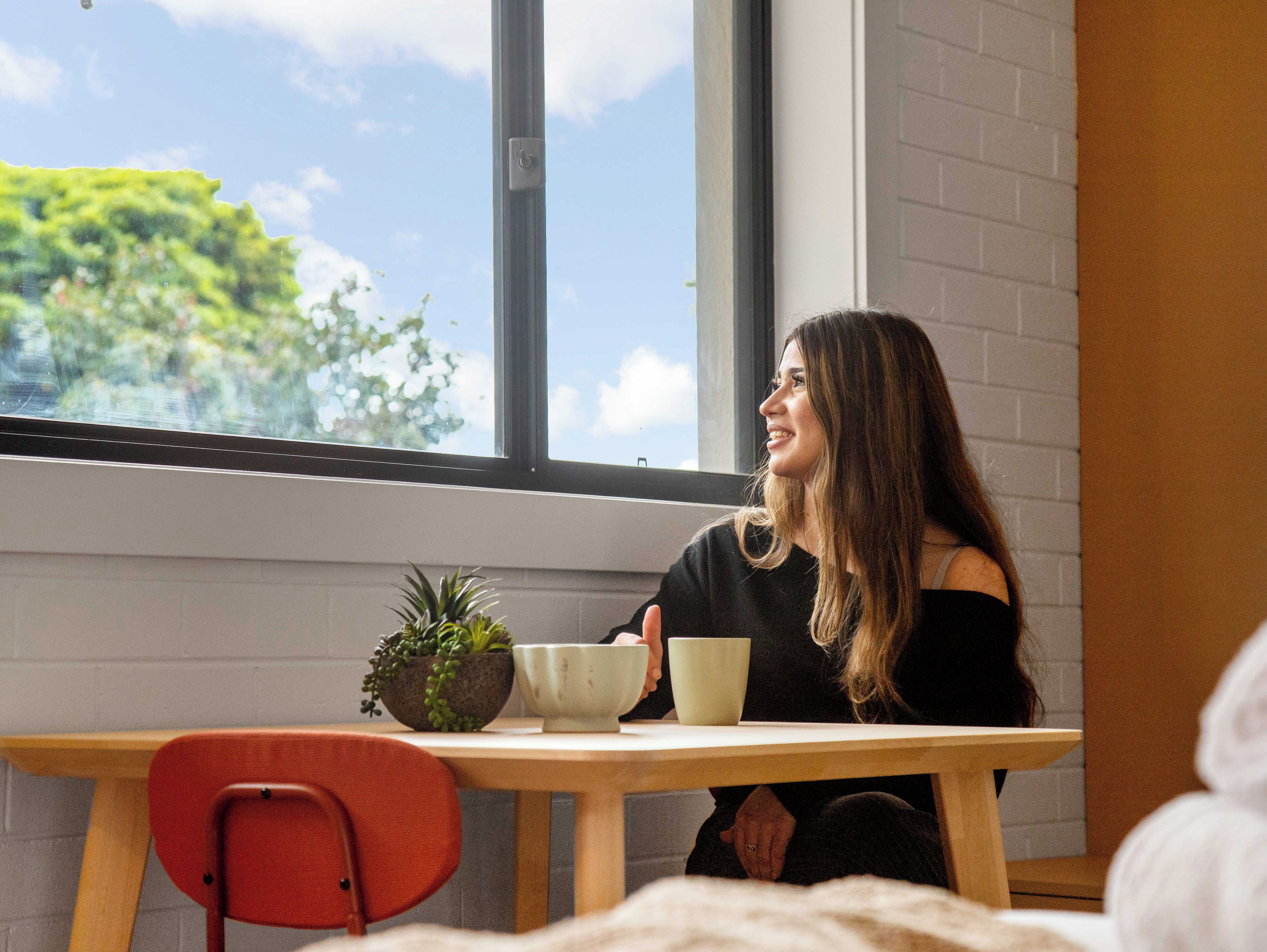 Una persona sentada a una mesa con vista al exterior, una planta en maceta y una taza.