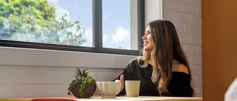 A person seated at a table with a window view, a potted plant, and a cup.