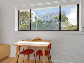 A room with a white brick wall, a window with a view of buildings and trees, a wooden table with two red chairs, and a small orange candle on the table.