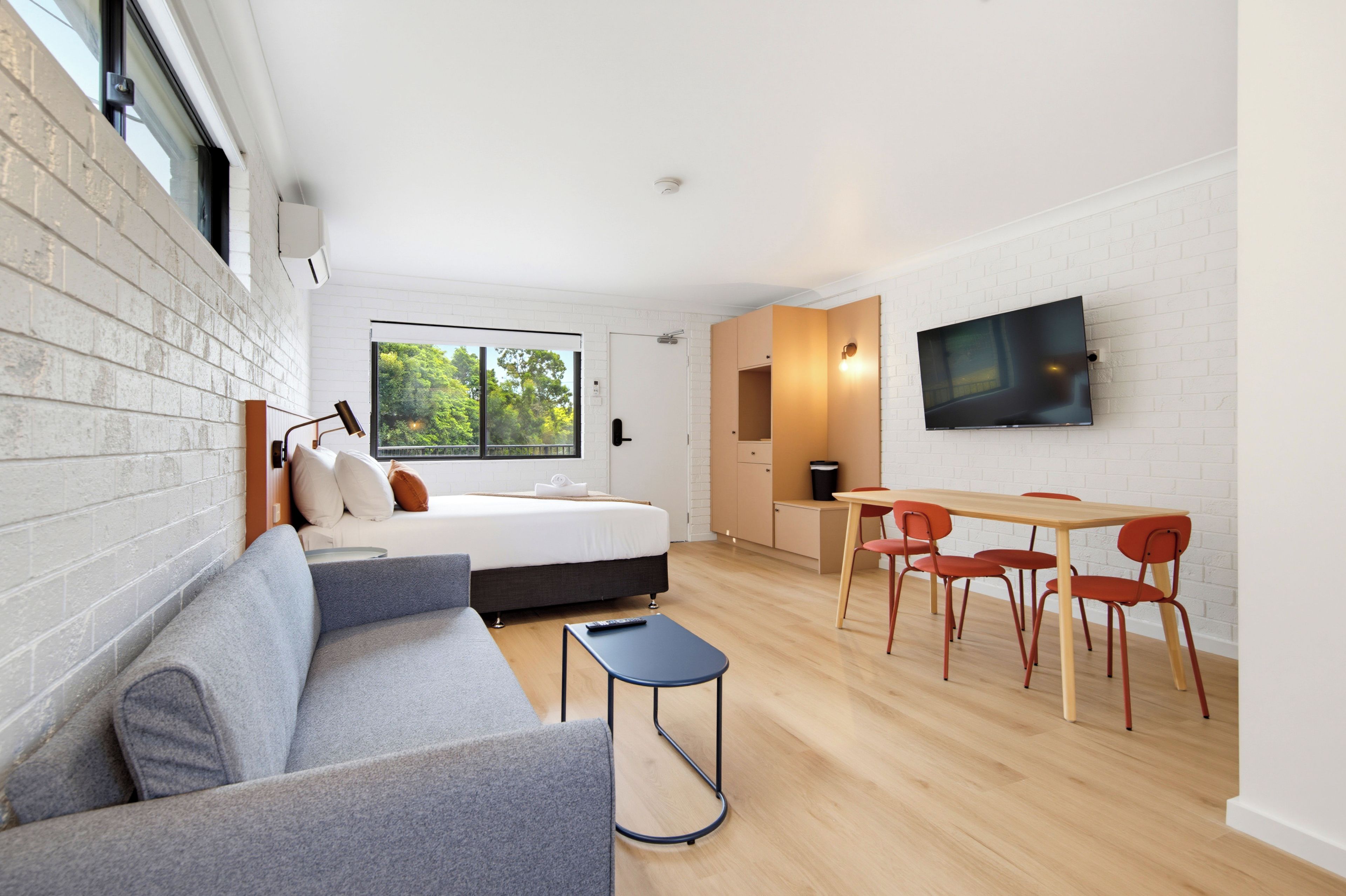 Una habitación de hotel moderna con una cama, un sofá, un comedor con sillas, un televisor montado en la pared y un ventanal grande.