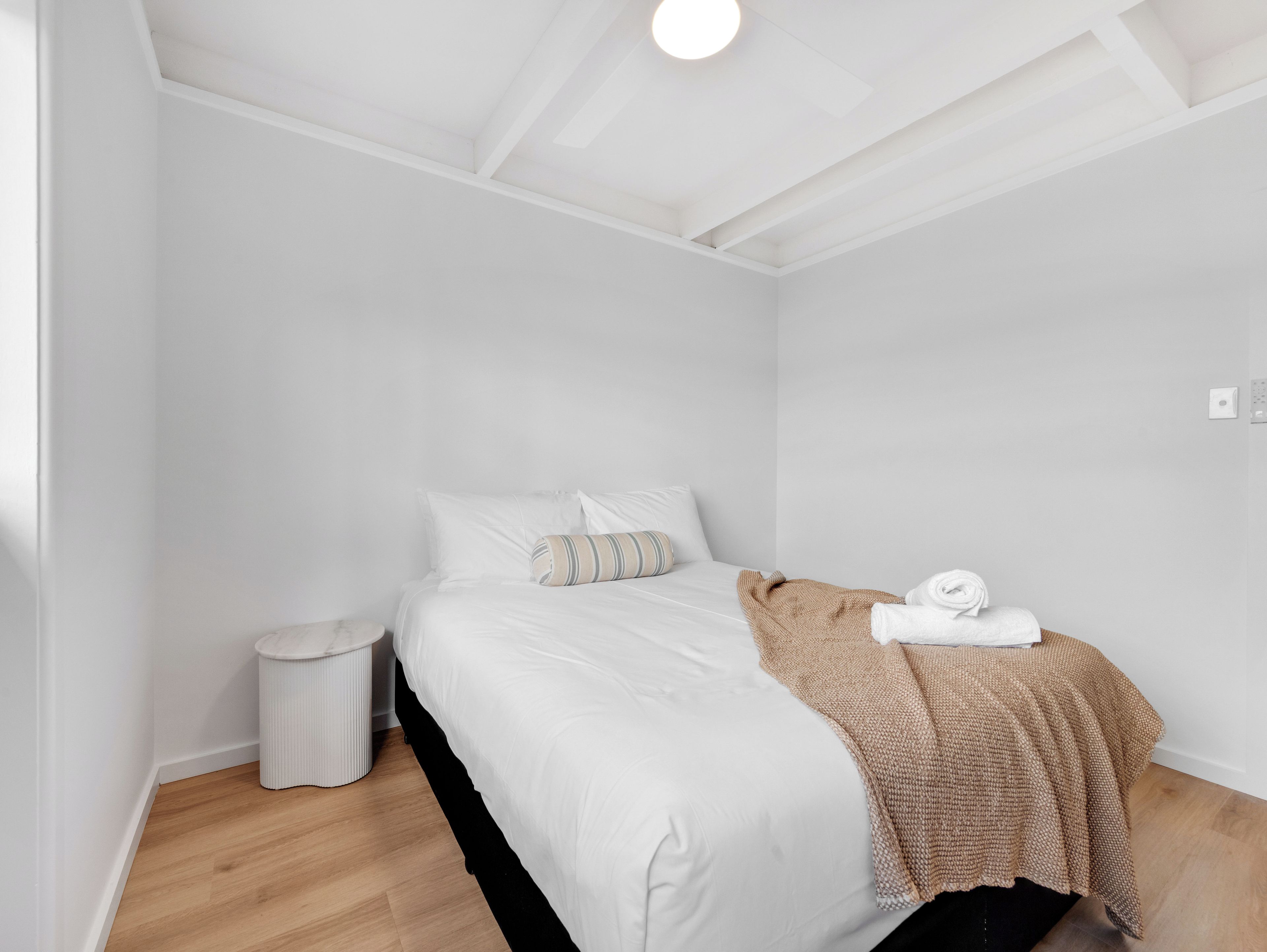 Un dormitorio minimalista con una cama individual, una mesita de noche pequeña y un cubo de basura.