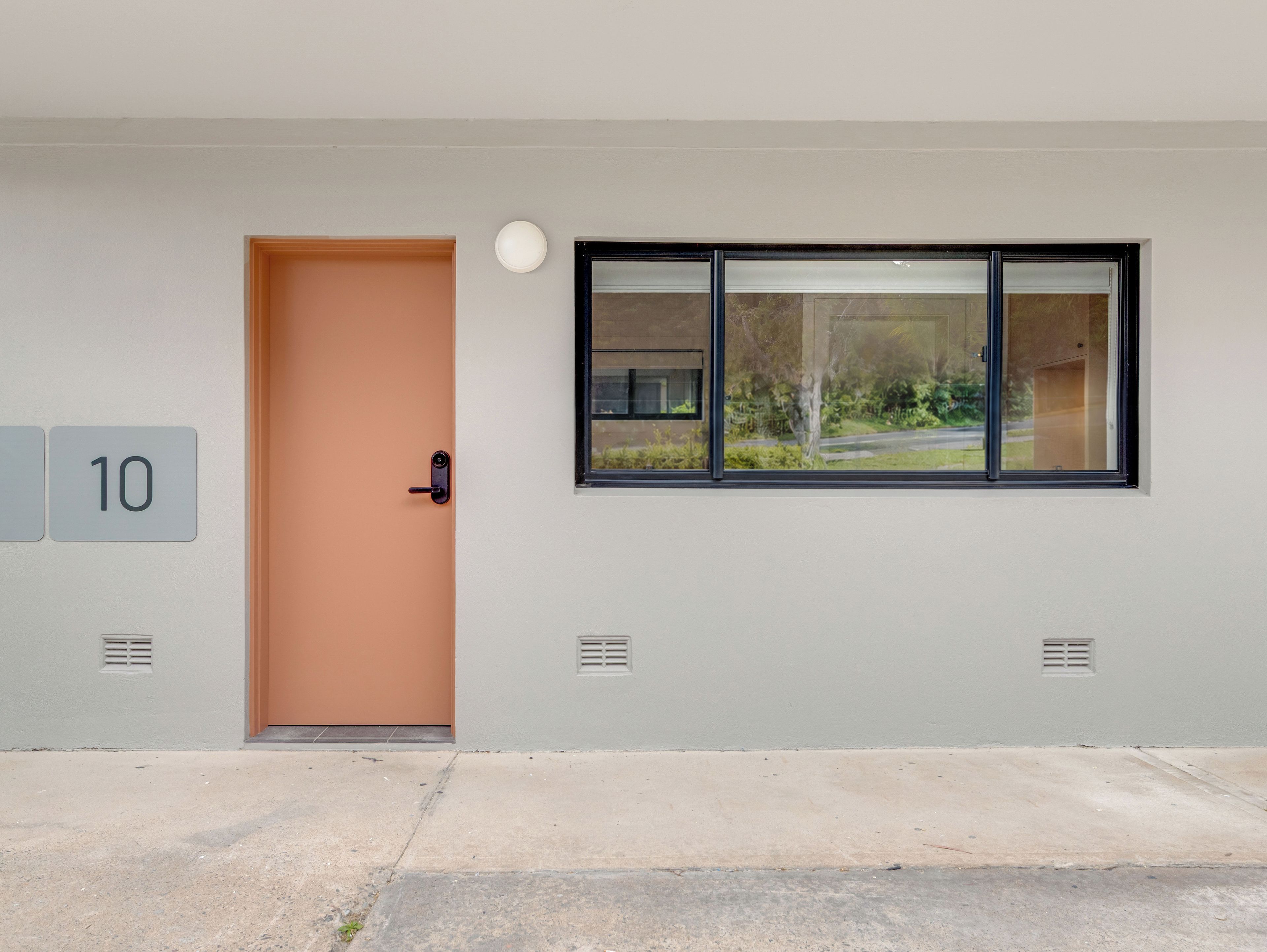 Entrada de un edificio con una puerta marcada '10', una ventana con vista a la vegetación y un accesorio de iluminación sobre la puerta.