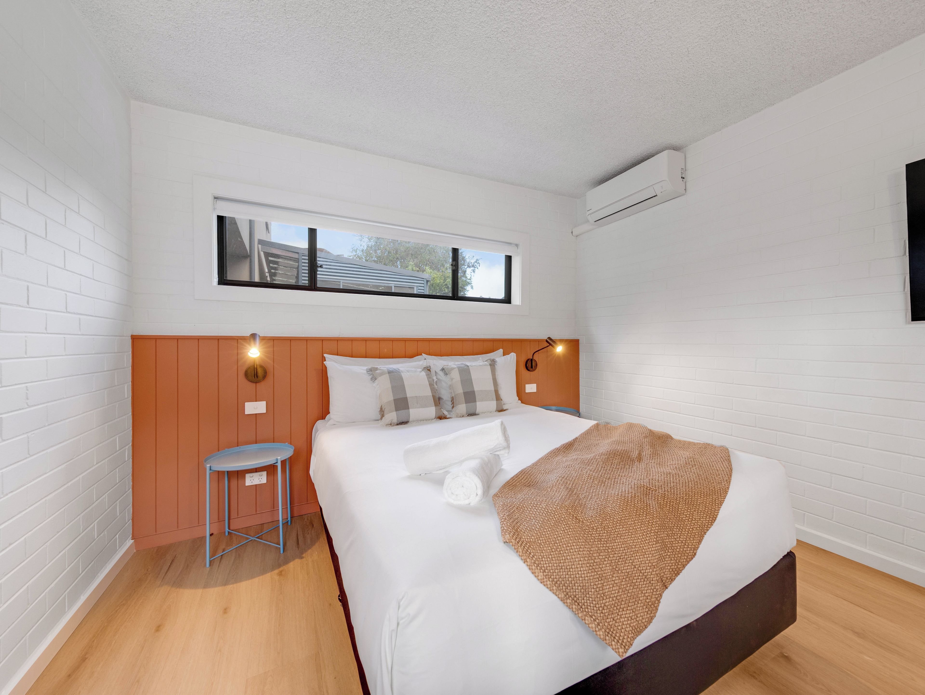 A bedroom with a bed, a window, a wall-mounted air conditioner, and a small stool.