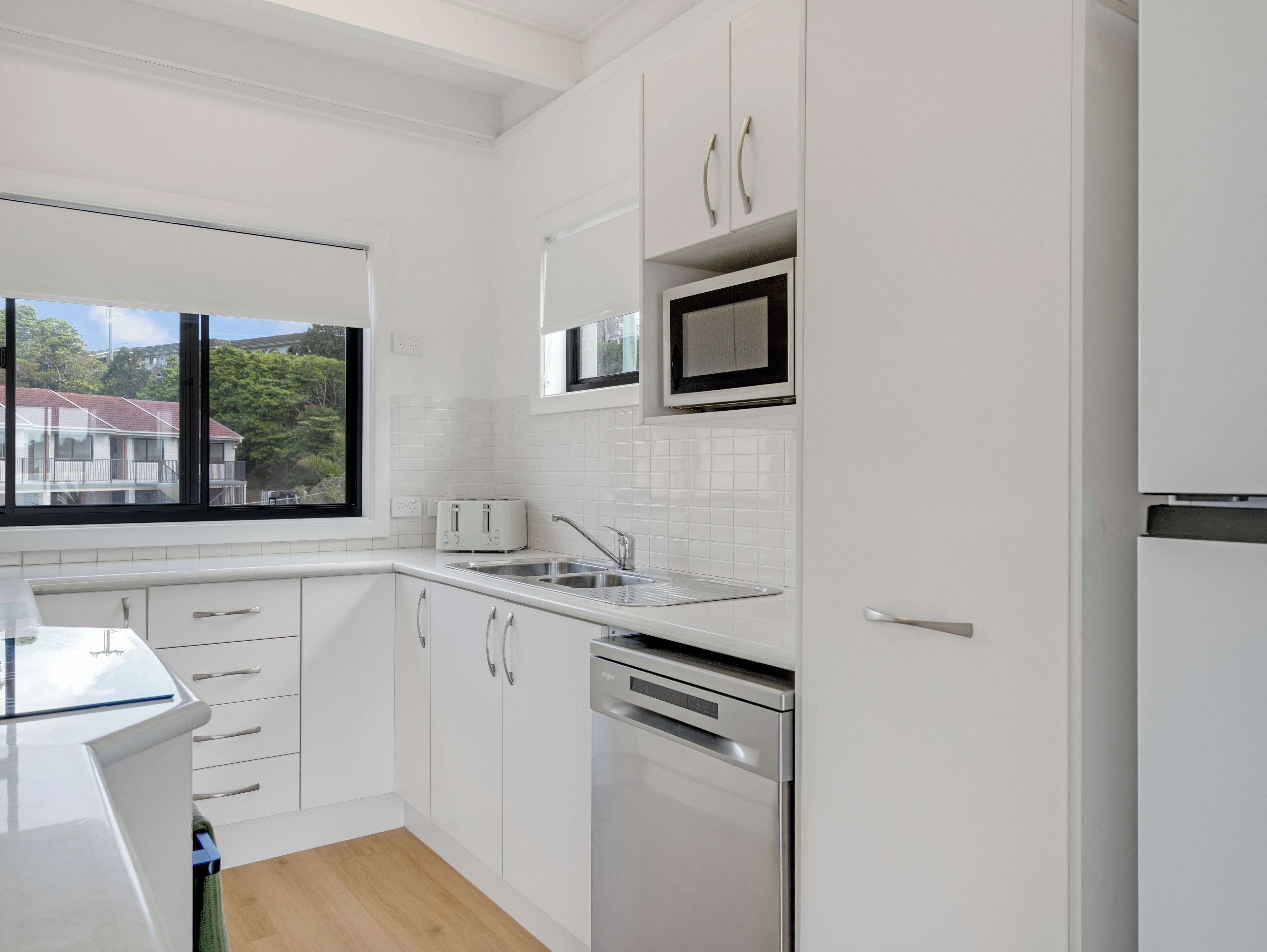 A modern kitchen with white cabinetry, a stainless steel dishwasher, and a window overlooking a residential area.