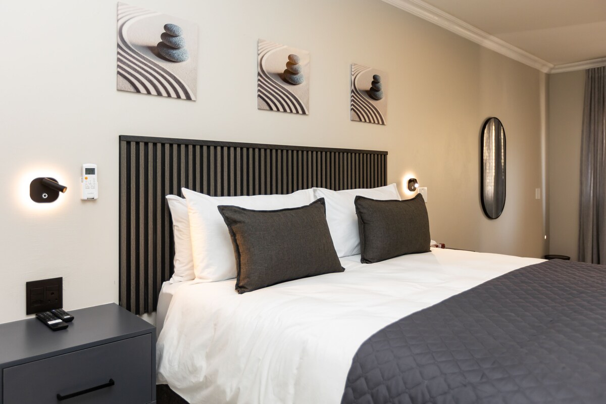 Una habitación de hotel moderna con una cama grande, una mesita de noche, un escritorio y un espejo montado en la pared.