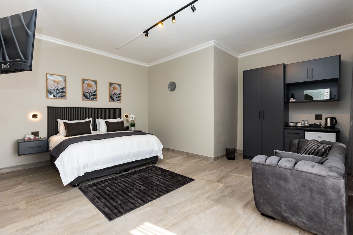 Un dormitorio moderno con cama, mesitas de noche, televisión de pantalla plana, reloj y una pequeña cocina.