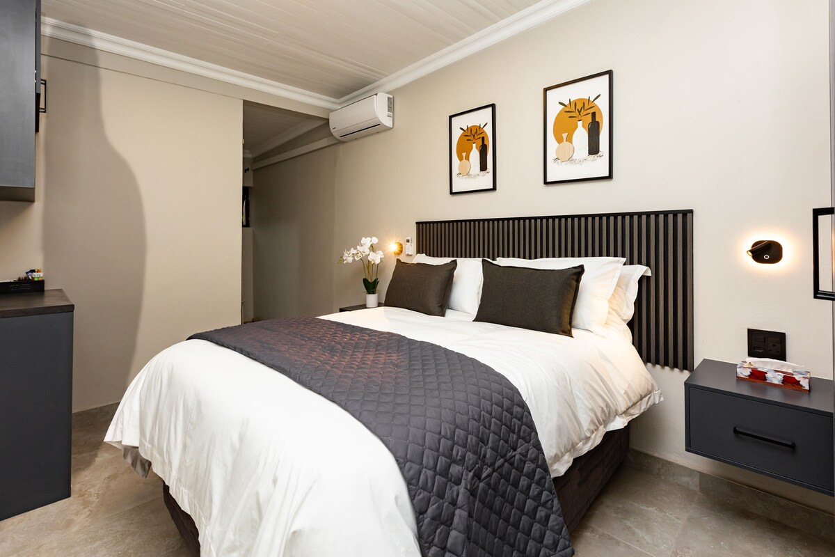 Un dormitorio moderno con una cama, mesitas de noche, un rincón de noche y dos obras de arte enmarcadas. Incluye aire acondicionado de pared.