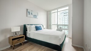 Un dormitorio con cama, una mesita de noche, una lámpara, una ventana con vistas a edificios y un cuadro en la pared.