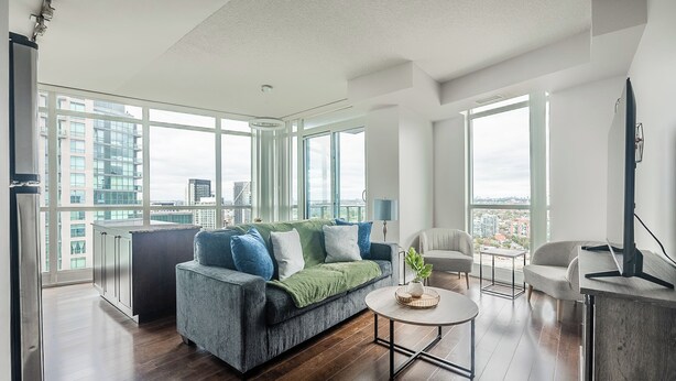 Una sala de estar moderna con sofá, mesa de centro y sillas, ventanas amplias con vistas a la ciudad y una zona de cocina.
