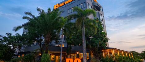 A modern hotel building with a prominent B2SURAT GRAND BOUTIQUE sign, surrounded by lush greenery and palm trees.
