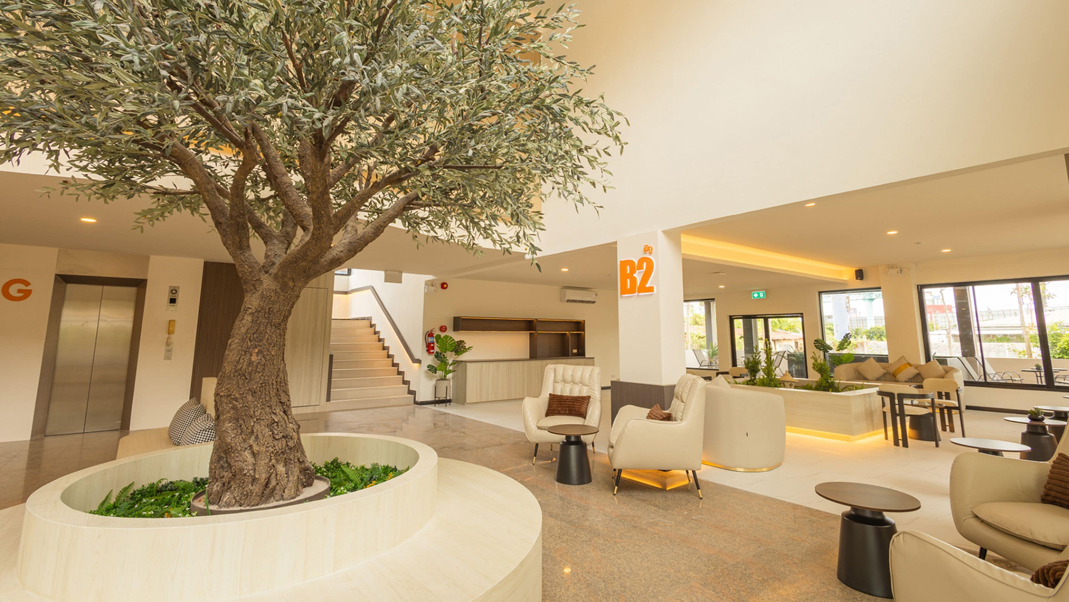 A modern lobby with a large tree, seating areas, a staircase, and a reception desk.
