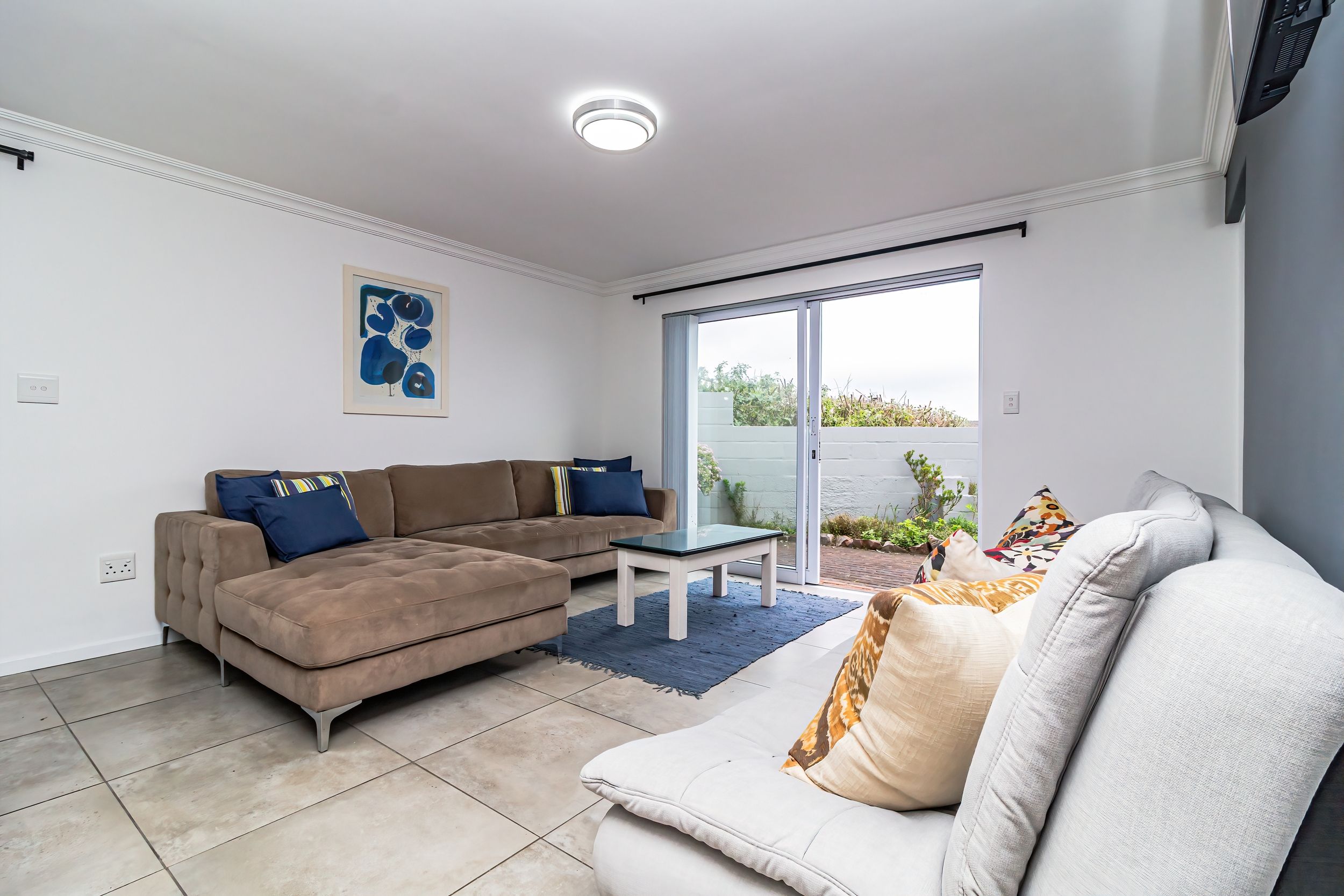 A living room with a brown sectional sofa, a white sofa, a small table, and a large sliding glass door leading to an outdoor area.