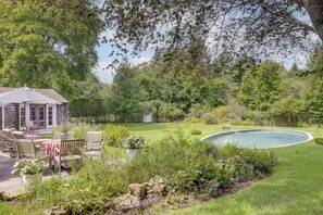Ein Pool, umgeben von einem üppigen Garten, einer Holzterrasse mit Stühlen und einem Pavillon.