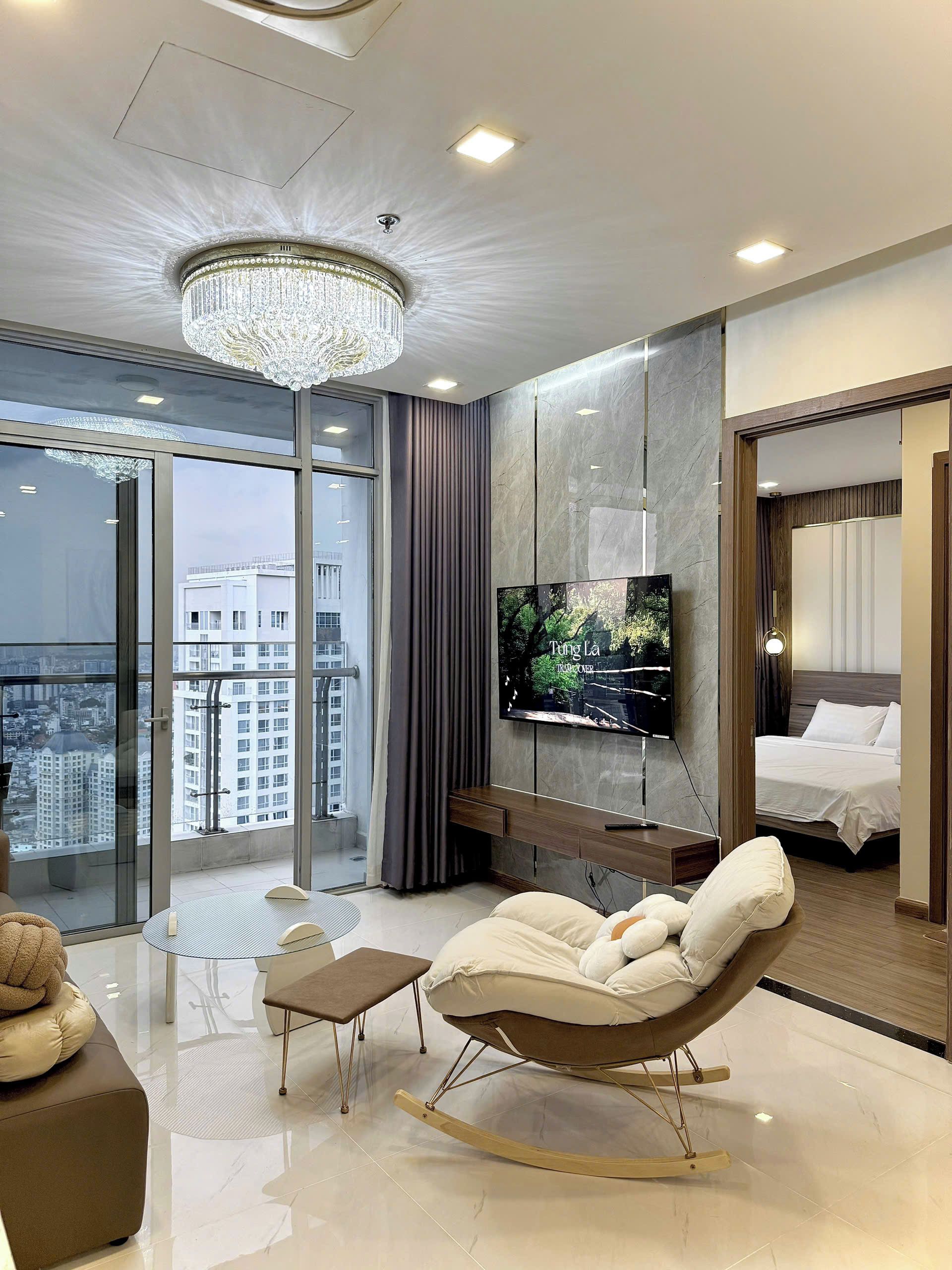 A modern hotel room with a large window, a chandelier, a flat-screen TV, a bed, and a seating area with a rocking chair.
