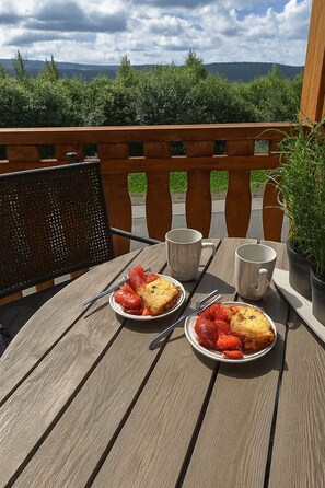 Ett träbord med två tallrikar mat och två koppar, placerat på en uteplats med utsikt över träd och kullar.