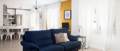 A living room with a sofa, a dining area with chairs, a kitchen with appliances, and a TV.