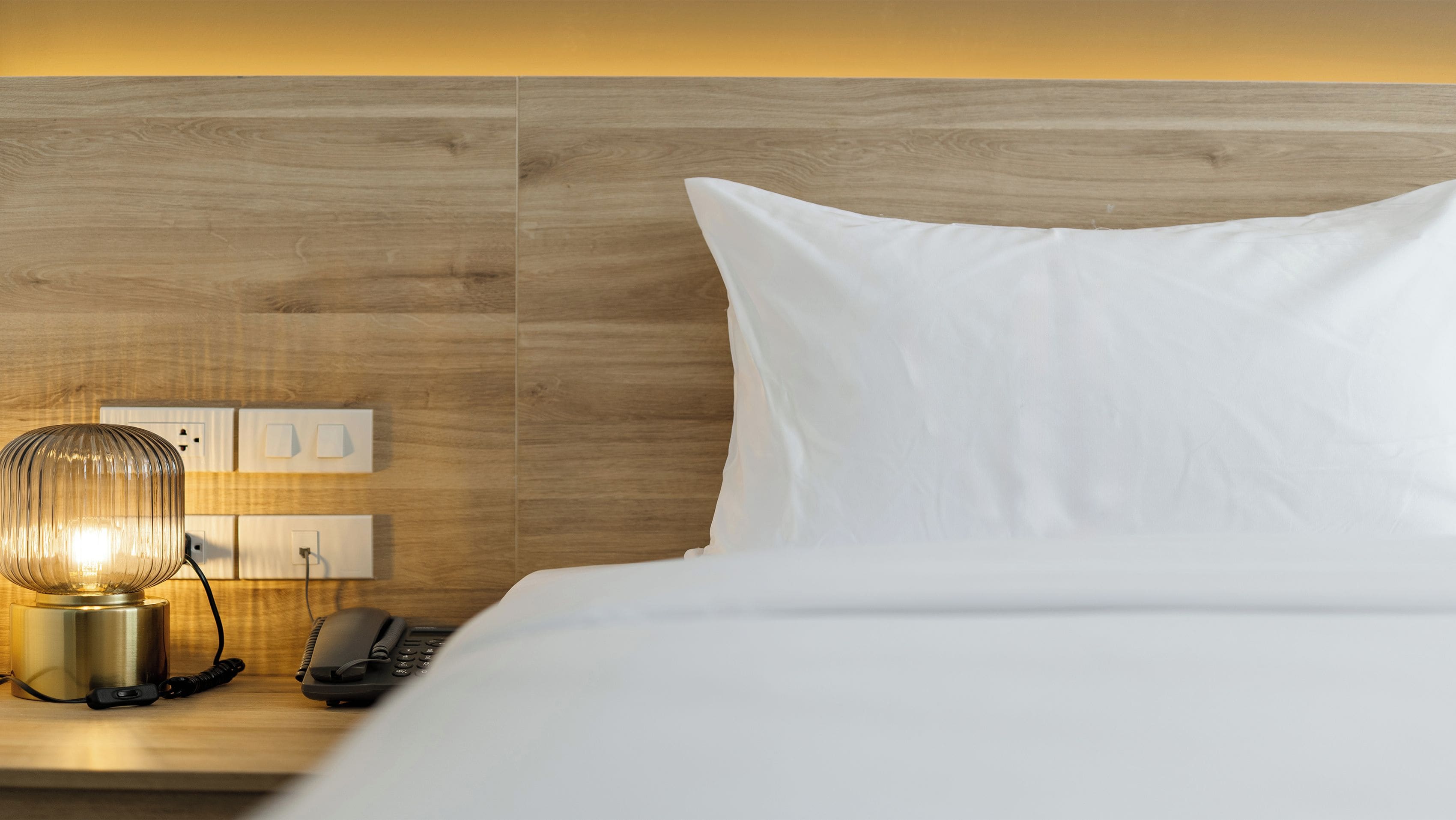 A hotel room with a wooden headboard, a bedside lamp, a telephone, and a white pillow.