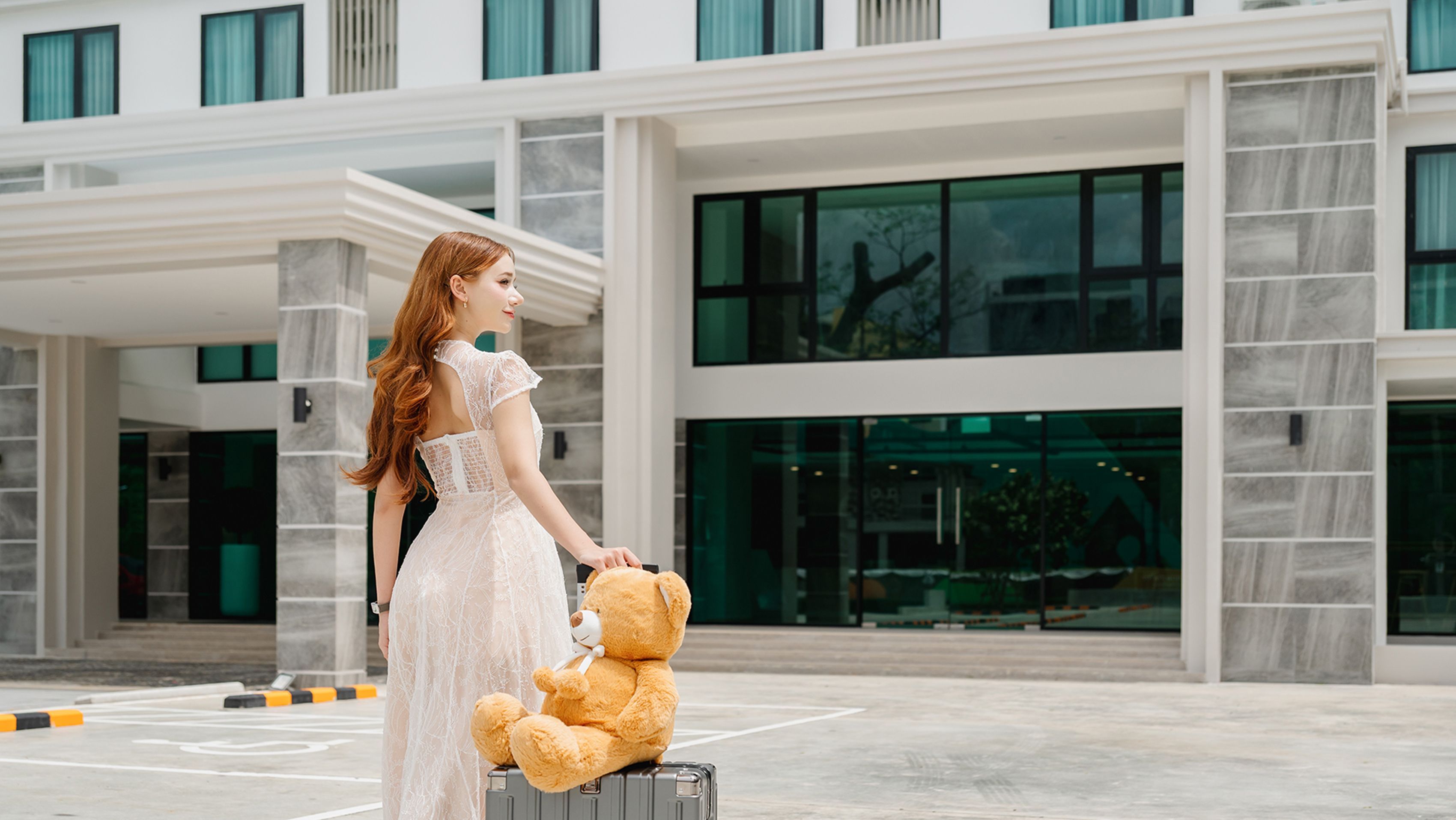 A person in a white dress holding a teddy bear and a suitcase in front of a modern building with large windows and a stone facade.