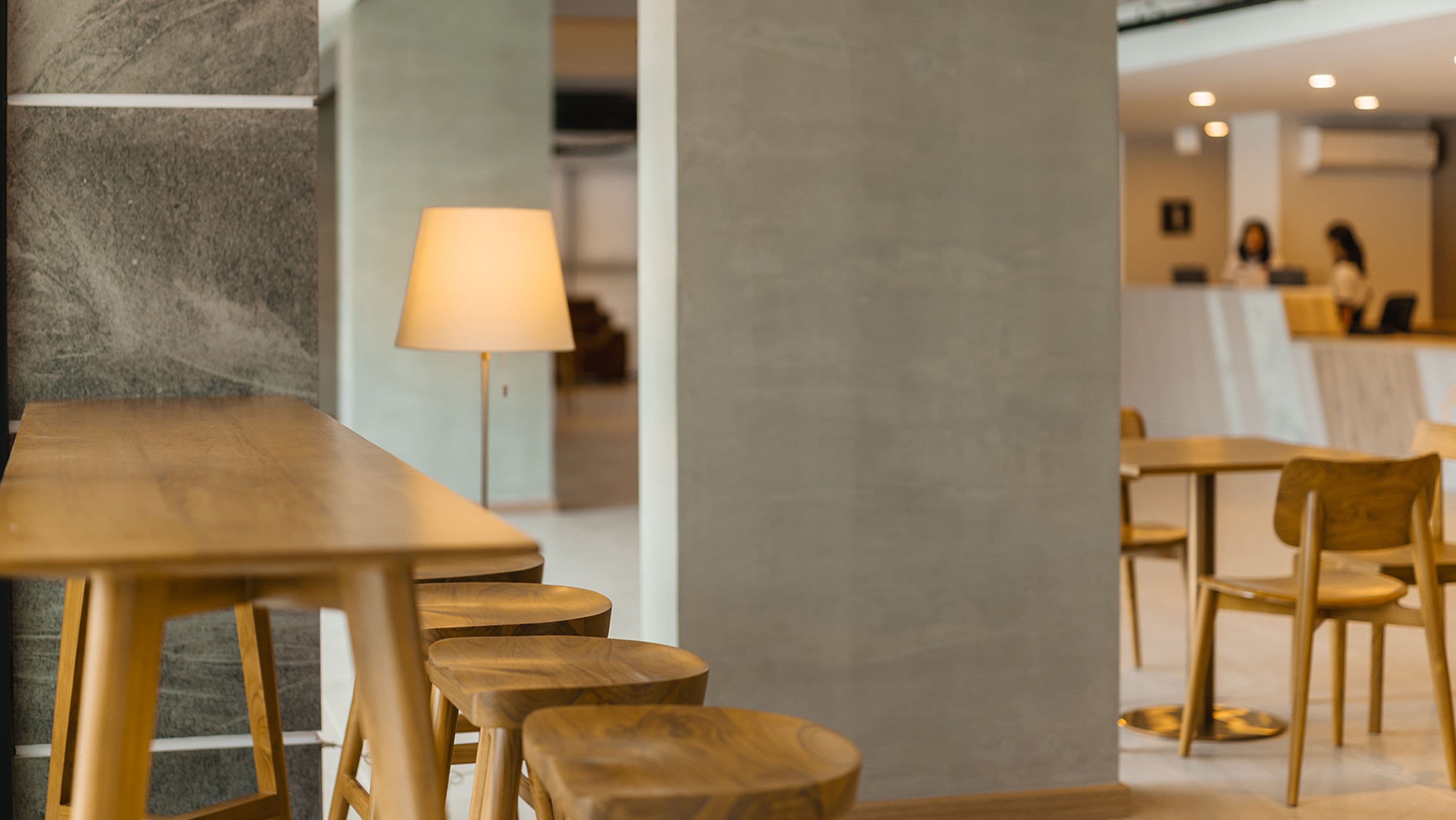 Un interior moderno con mesas y sillas de madera, una lámpara y una zona de recepción al fondo.