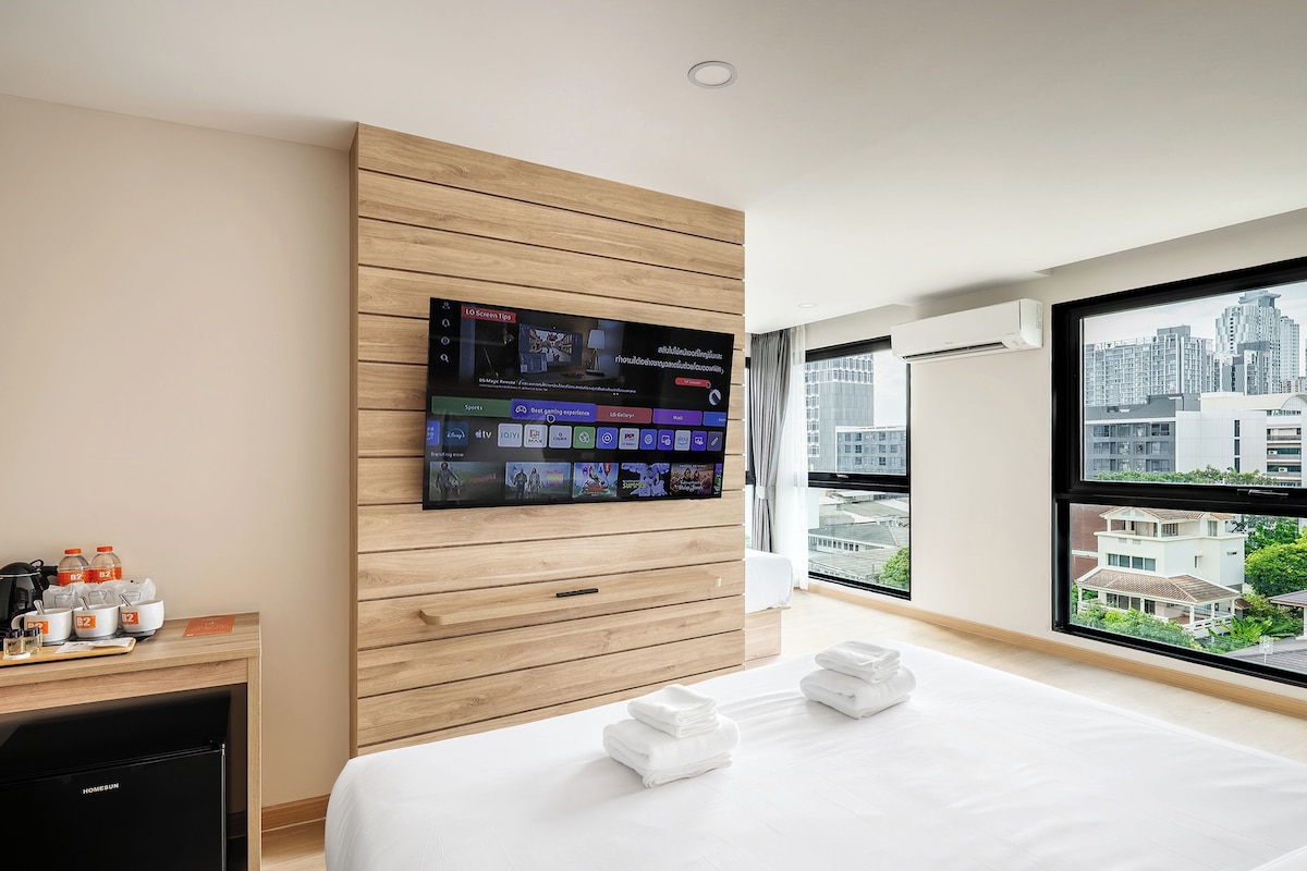 Une chambre d’hôtel dotée d’un grand téléviseur fixé sur un mur en panneaux de bois, d’un lit avec des serviettes soigneusement pliées, d’une petite kitchenette équipée d’une machine à café et offrant une vue sur les bâtiments de la ville par de grandes fenêtres.