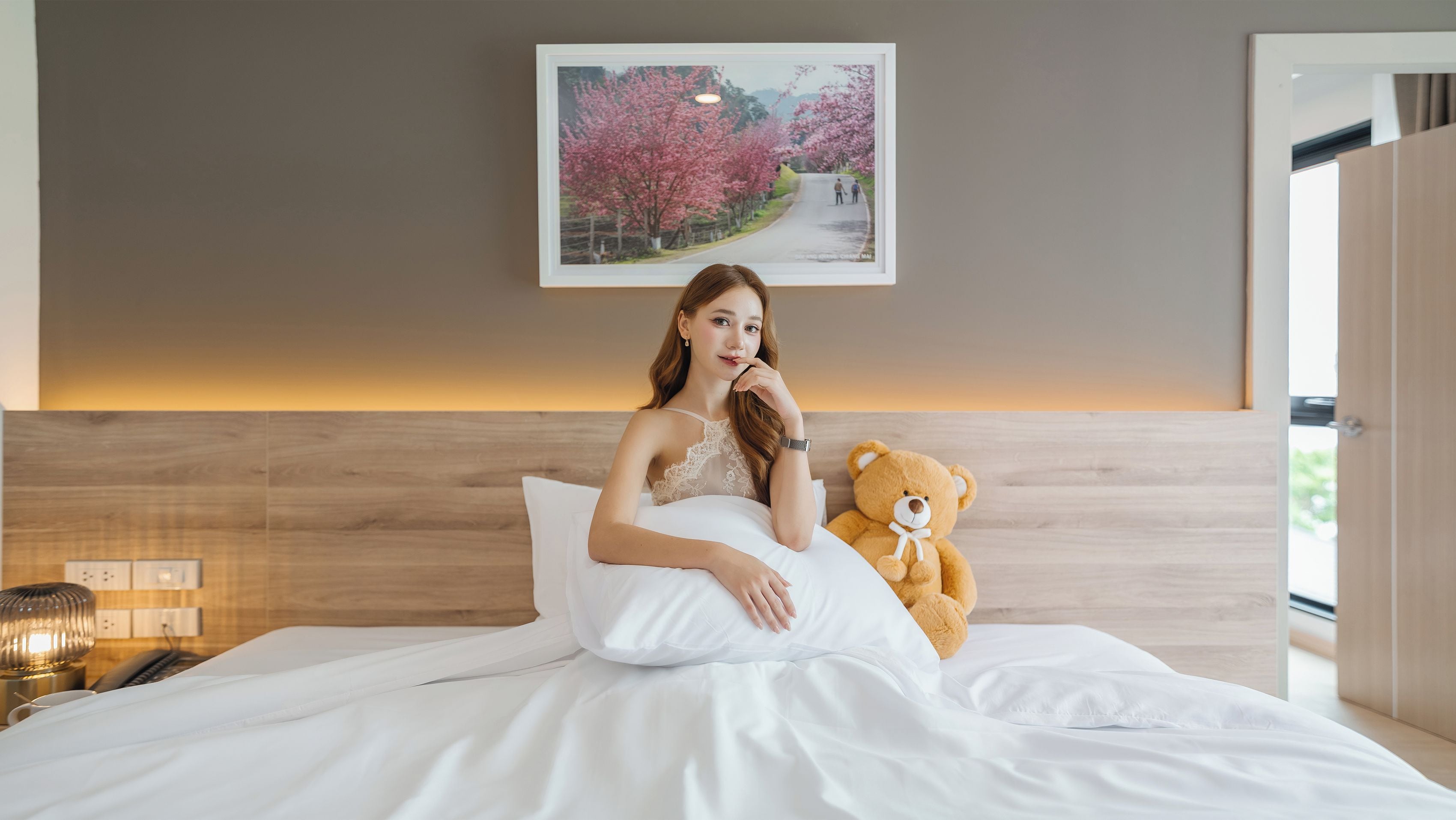 A person sitting on a bed with a teddy bear beside them, a framed picture on the wall, and a bedside lamp.