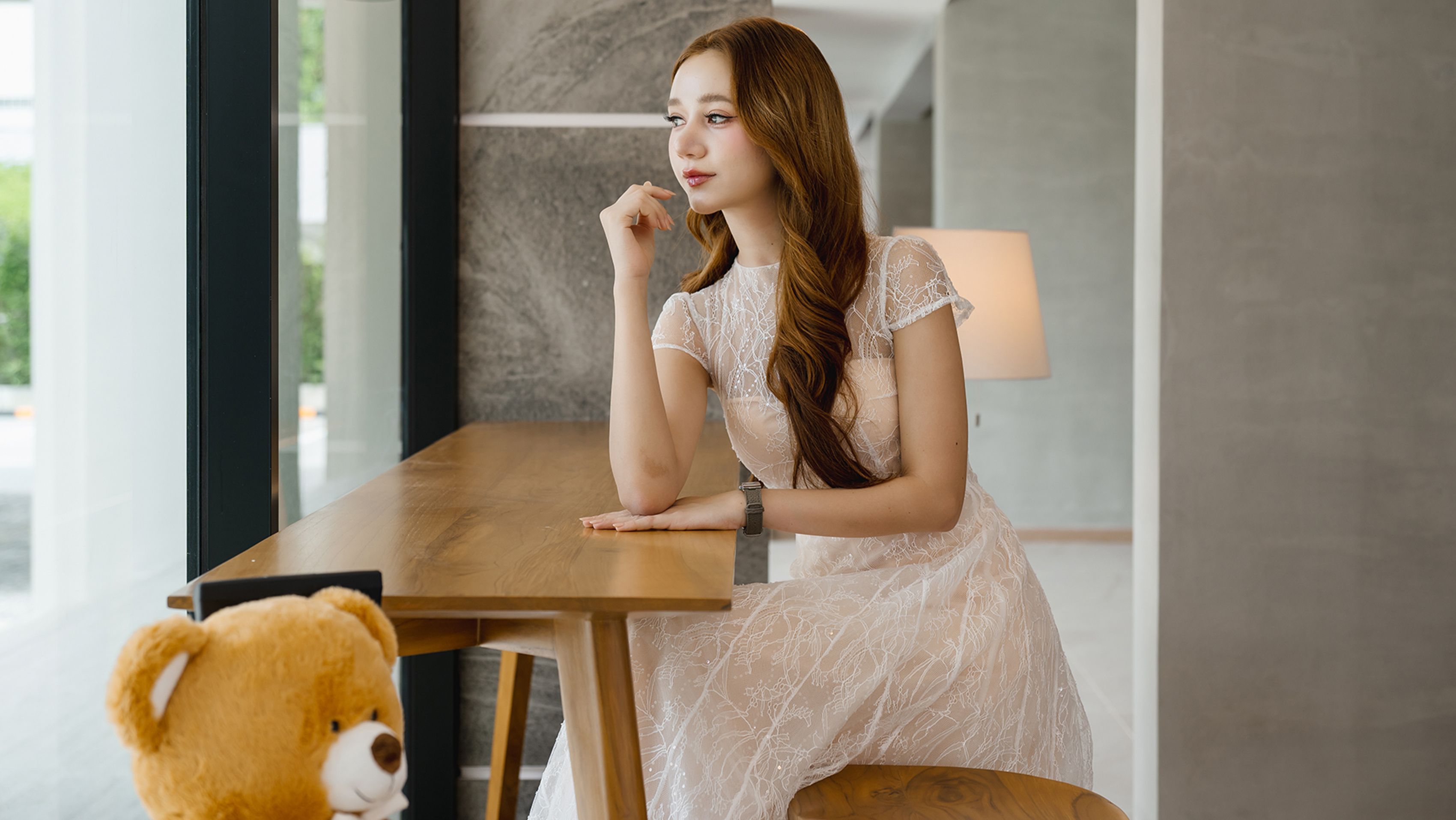 A person seated at a wooden table in a room with a large window and a lamp.