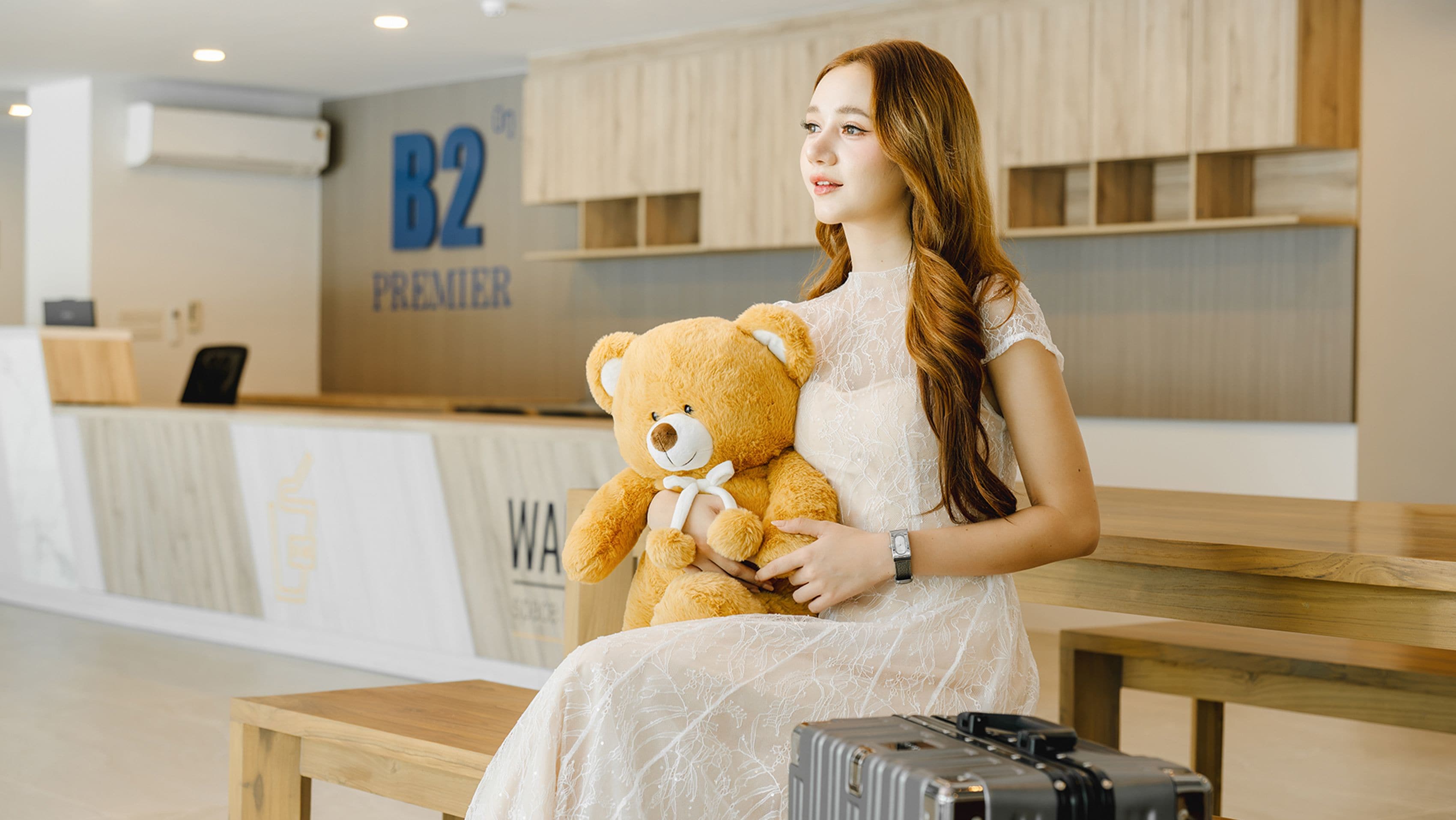 A woman sitting on a bench in a hotel lobby with a teddy bear, holding a smartphone, and a suitcase beside her.