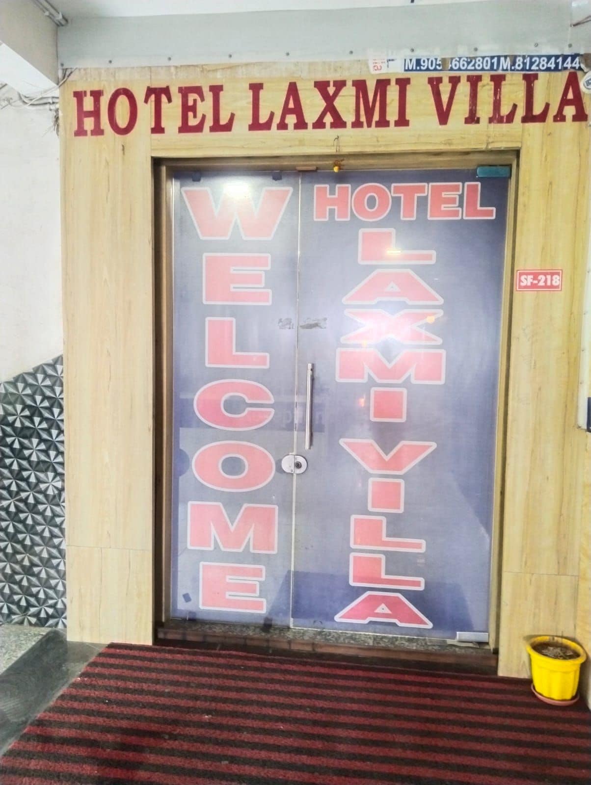 Entrada de un hotel con fachada de madera y puerta de cristal. Se exhiben claramente las palabras 'WELCOME' y 'HOTEL LAXMI VILLA' en el cristal.