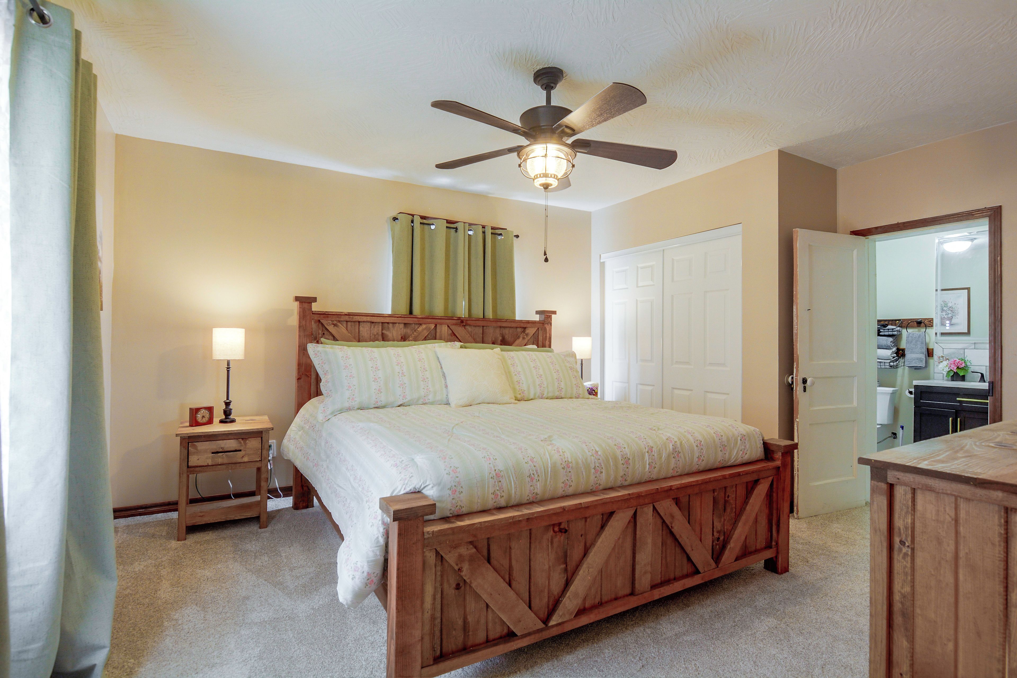 Quarto com cama de madeira, mesa de cabeceira com luminária, ventilador de teto e porta para um banheiro.