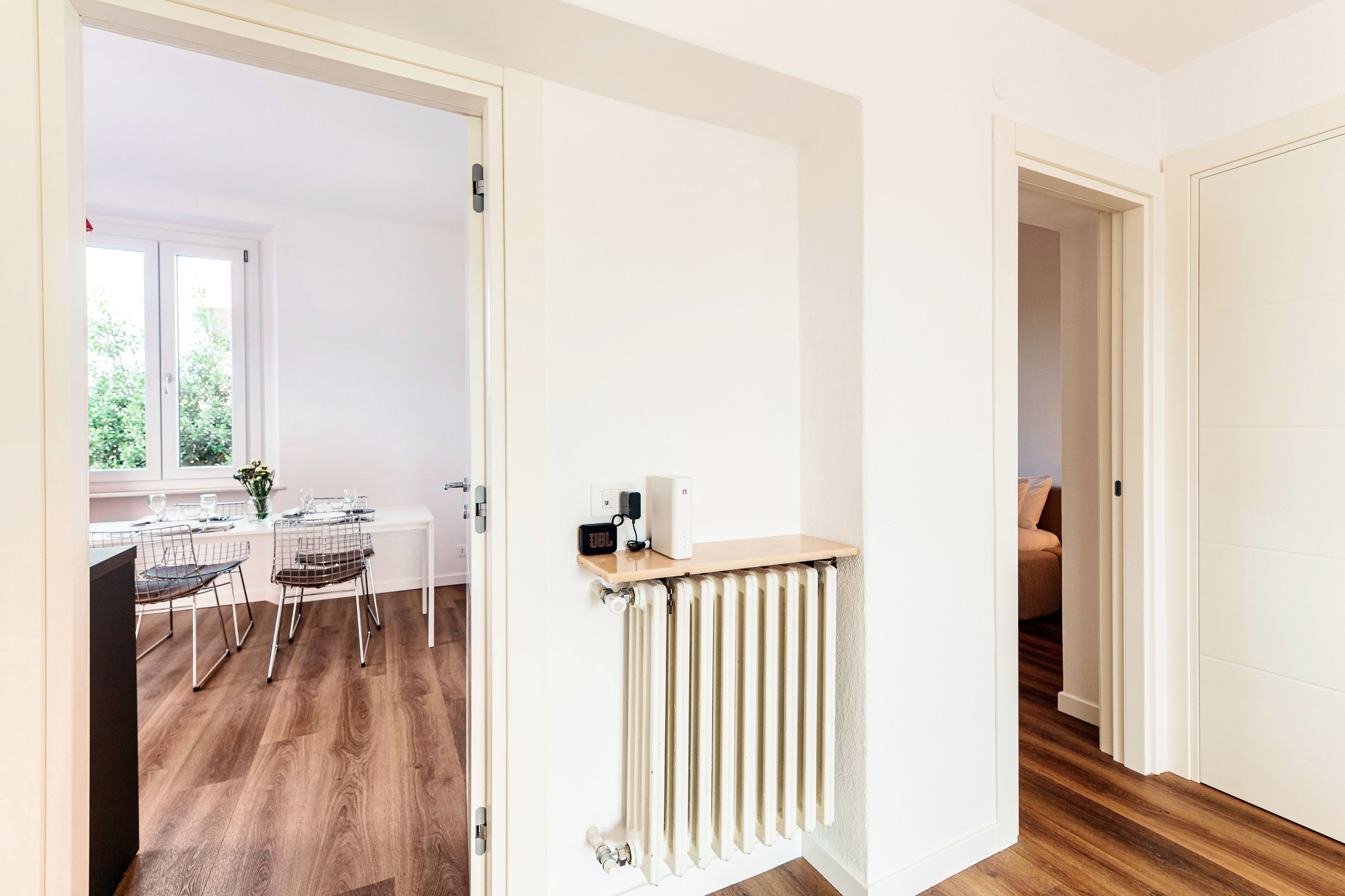 A hallway with a radiator, a door leading to a dining area with a table and chairs, and a view of a window with greenery outside.