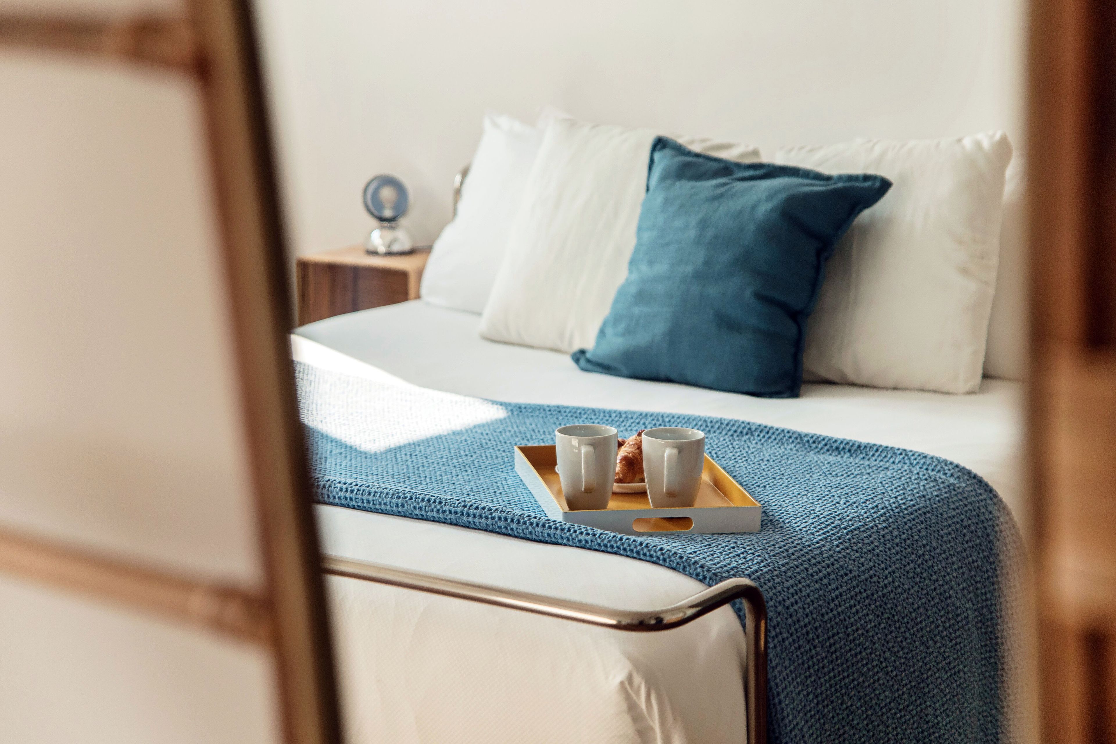 A neatly made bed with a blue blanket, two white pillows, and a tray with two cups and a small plate.