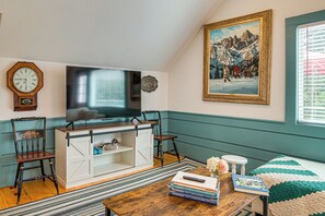 A living room with a television, a clock, a painting, and a bookshelf. - Walk to Main St! Osterville Cottage w/ Patio (Osterville)