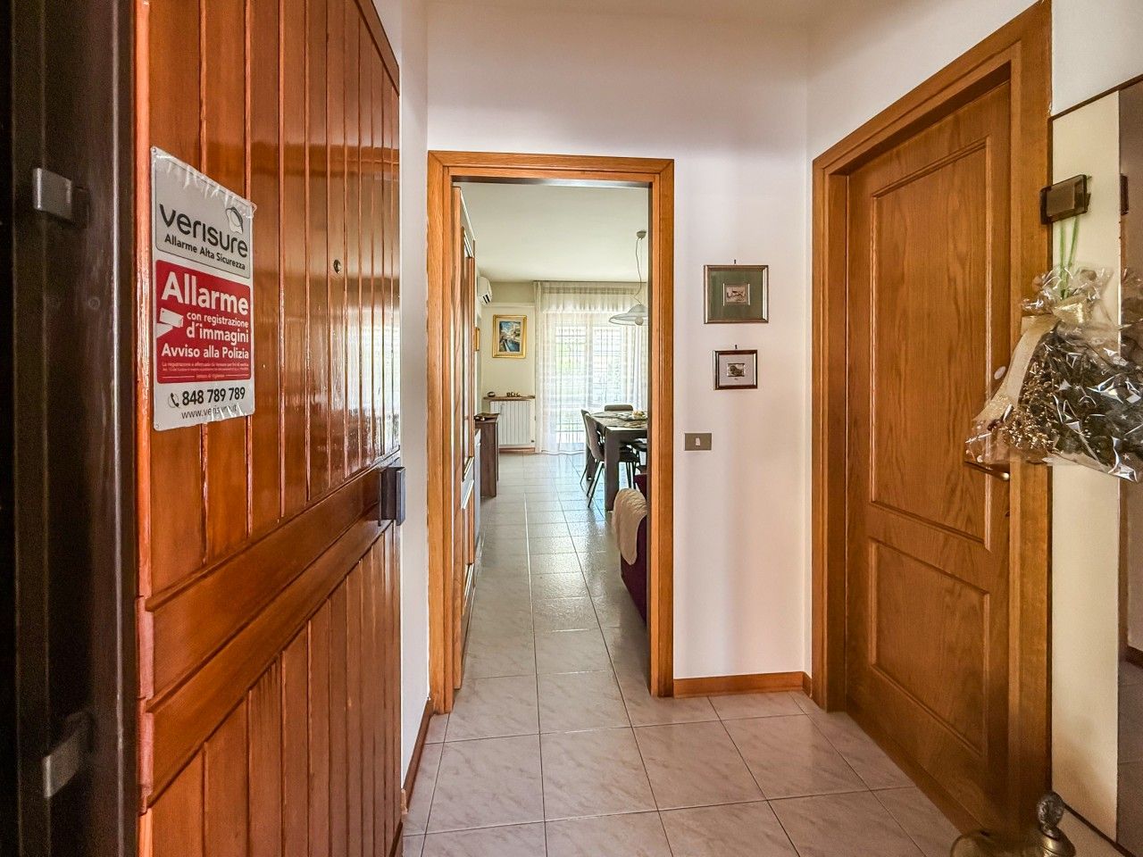 A hallway with wooden doors, a sign for Allarme security service, and a dining area in the background.