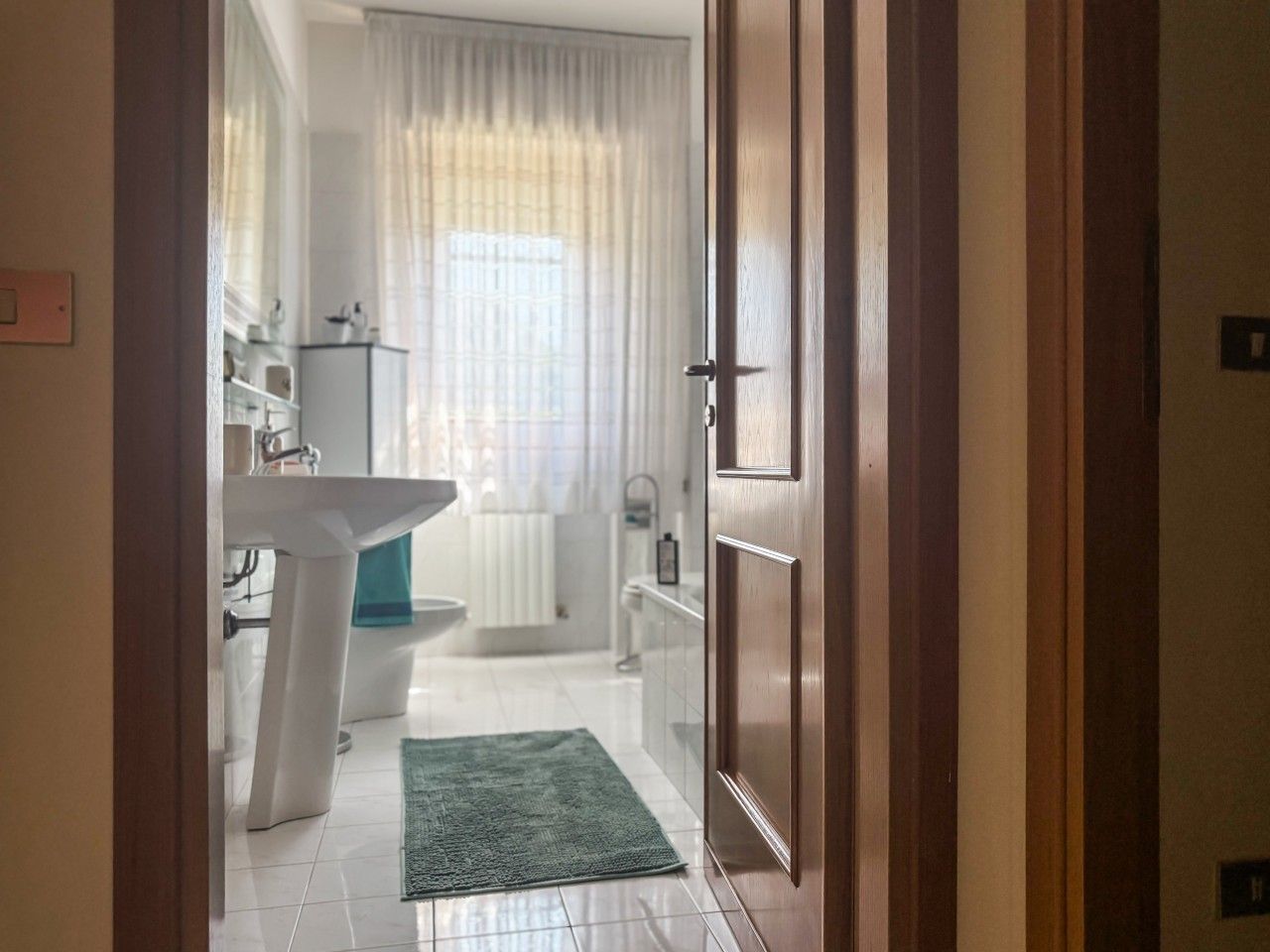 A bathroom with a white sink, toilet, and bathtub, a green rug, and a window with curtains.