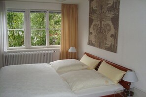 Een hotelkamer met een bed, een raam met gordijnen, een radiator en een wandlamp.