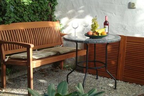 Een houten bank met een bekleed zitvlak, een ronde tafel met een metalen onderstel, en een schaal fruit met een fles wijn.