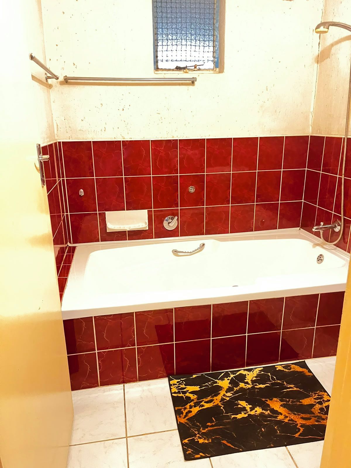 Un baño con una bañera blanca, paredes con azulejos rojos, un ventanal pequeño y un tapete con estampado negro y dorado en el suelo.