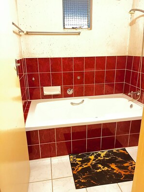 A bathroom with a white bathtub, red tiled walls, a small window, and a black and gold patterned rug on the floor.