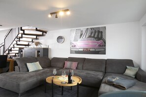 A modern living room with a grey sectional sofa, a wooden coffee table with wine glasses, a clock, and a cityscape painting. - House Relax by the lake (Göhren-Lebbin)