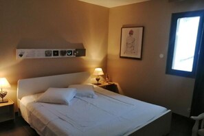 A hotel room with a bed, a nightstand with a lamp, a wall-mounted shelf with pictures, and a window. - Holiday home in Provence (Merindol)