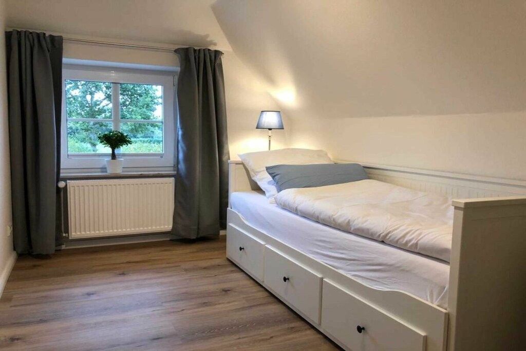 Ein Einzelzimmer mit Fenster, Heizkörper, Nachttischlampe und einer Pflanze auf dem Fensterbrett.