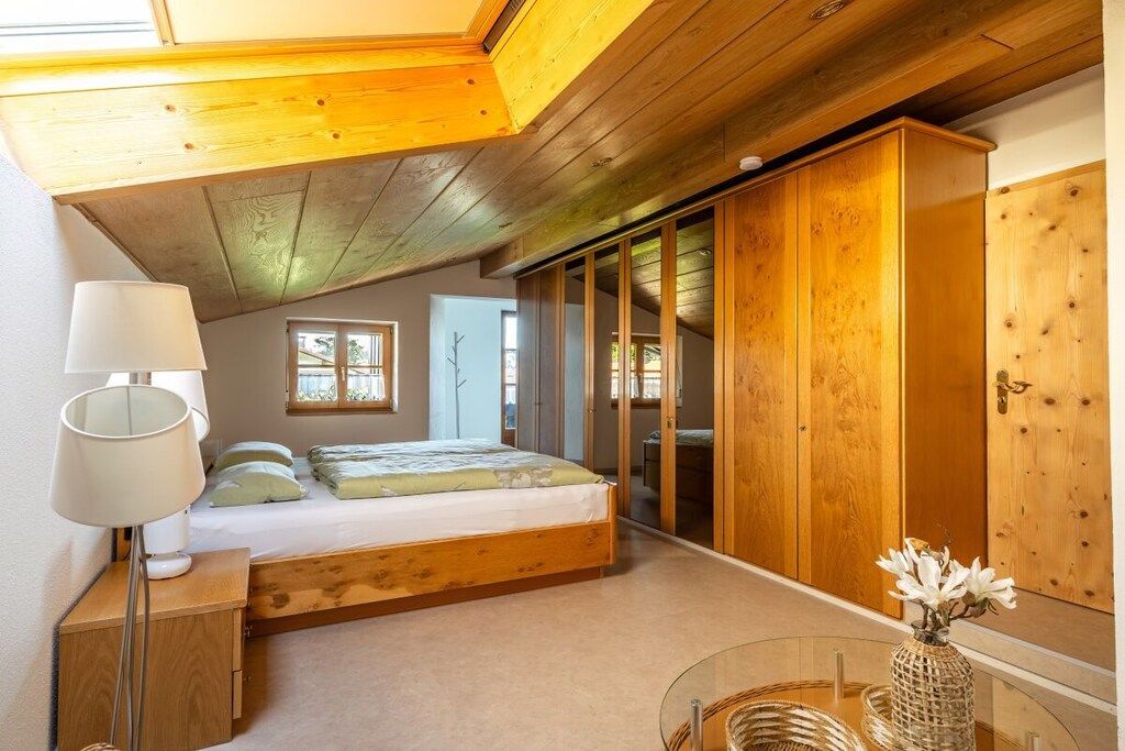 A bedroom with a bed, bedside lamp, wooden wardrobe, and a small table with a vase.