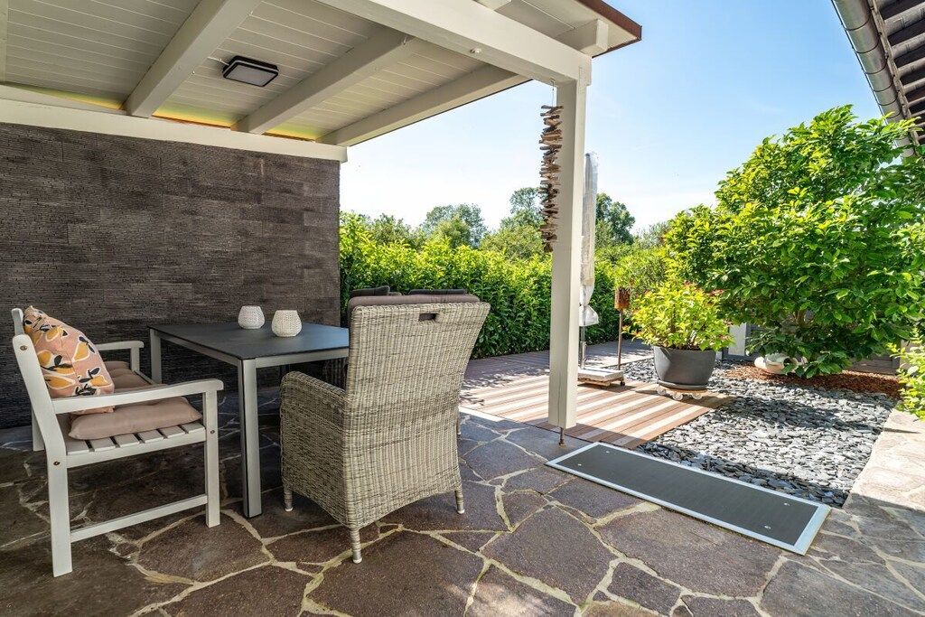 Eine Terrasse im Freien mit Tisch und Stühlen, einem Steinboden und einem überdachten Bereich mit Blick auf Grünflächen.